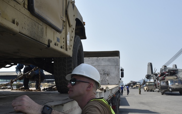 A worker wearing a white hard hat, yellow safety vest, sunglasses, and a watch is inspecting or overseeing the loading/securing of a military HMMWV (Humvee) onto a flatbed trailer at what appears to be a port or logistics staging area. In the background, military helicopters including what appear to be UH-60 Black Hawks and a CH-47 Chinook are visible on the tarmac. Multiple personnel and port infrastructure are visible in the background.