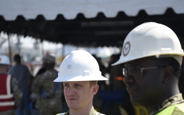 Two U.S. Navy personnel in camouflage uniforms wearing white hard hats and high-visibility yellow safety vests are engaged in conversation at what appears to be a port or construction site. The foreground subjects are in sharp focus while additional military and civilian personnel are visible in the blurred background. The setting suggests a logistics, humanitarian, or construction operation.