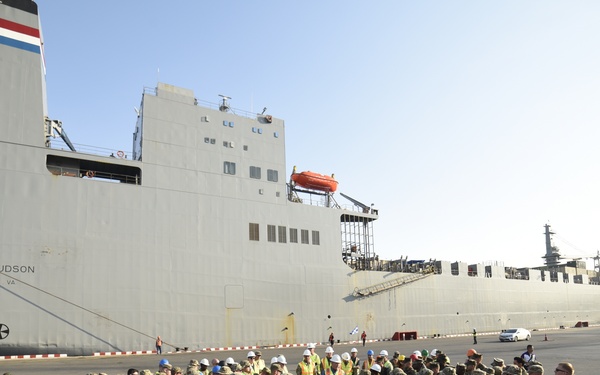 A large group of military personnel in camouflage uniforms are assembled on a dock beside a large cargo/military vessel named 'Cape Hudson' homeported in Norfolk, VA. The personnel are wearing blue hard hats and high-visibility yellow vests, suggesting a port loading or briefing operation. The scene appears to be a military embarkation or debarkation event at a naval port facility.