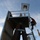 A worker in safety gear is climbing a metal ladder attached to an industrial tower or navigation aid structure. The structure features a red and white warning light at the top, a solar panel, and large rusted support columns. A coiled rope hangs freely from the structure near the worker.