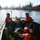 Two personnel in safety gear are aboard a metal work boat traveling along a calm river or waterway. Both individuals are wearing orange immersion/survival suits, helmets, and protective eyewear, suggesting a water rescue, environmental monitoring, or emergency response operation. The surrounding landscape features coniferous and deciduous trees along the riverbanks under clear winter conditions.