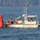 A U.S. Coast Guard vessel is positioned alongside a large red navigational buoy on open water. Crew members dressed in orange survival suits are actively working on or near the buoy, likely performing maintenance or repositioning operations. A wooded hillside is visible in the background under overcast conditions.