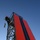A worker equipped with safety harness and helmet is positioned at the top of a metal lattice tower structure, appearing to rappel or perform maintenance work. The tower supports a large billboard or display panel featuring bold red and dark navy/black vertical stripes. The image is taken from a low angle looking upward against a clear blue sky.