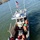 An aerial view of a U.S. Coast Guard response boat on open water, with multiple crew members in orange immersion suits and safety helmets visible on deck. The vessel is equipped with an American flag, blue emergency lights, radar dome, and various rescue equipment. Crew members appear to be conducting or preparing for a search and rescue or emergency response operation.