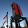 Workers in safety gear are performing maintenance on a large navigational dayboard marker mounted on a steel structure over or near a waterway. The scene is photographed from the deck of a work vessel, with two American flags visible and equipment scattered on the boat deck. The large red and black rectangular dayboard is a channel marker used for maritime navigation.