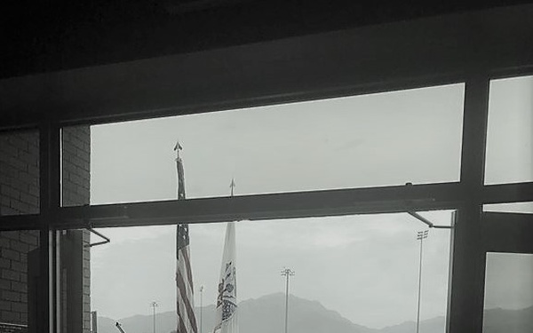 A black-and-white photograph taken from inside a building looking through glass doors at a military ceremony or formation taking place outdoors on a wet surface, suggesting recent rainfall. A color guard or honor guard is visible holding the American flag and what appears to be a unit guidon or organizational flag. Mountains are visible in the background, and the overcast sky contributes to the somber tone of the image.