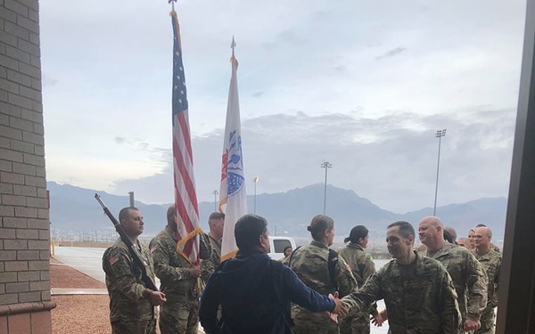 A civilian in dark civilian clothing is greeting a formation of U.S. Army soldiers in OCP (Operational Camouflage Pattern) uniforms outside a military building. The scene appears to be a formal reception or welcome ceremony, evidenced by a color guard holding the U.S. flag and a unit guidon. The setting appears to be a military installation with mountains visible in the background under overcast skies.