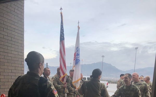 A group of military personnel in U.S. Army camouflage uniforms (OCP pattern) are gathered on a wet tarmac or paved area, appearing to participate in a welcome or greeting ceremony. Soldiers are shaking hands in what appears to be a receiving line formation, with color guard members holding the U.S. flag and a unit guidon in the background. The setting appears to be a military installation with mountains visible in the background under overcast skies.
