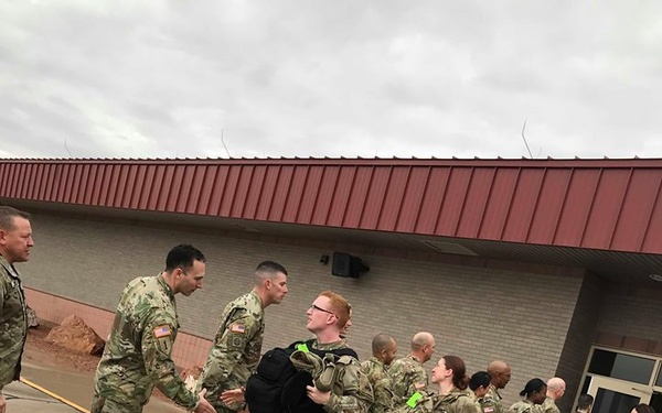 A group of U.S. Army soldiers in OCP (Operational Camouflage Pattern) uniforms are lined up on a wet outdoor tarmac or parking area, appearing to go through a reception or greeting line with senior personnel. Some soldiers are carrying tactical backpacks and have colored tags (green) attached to their gear, suggesting a processing or arrival/departure event. Two civilians are visible on the right side of the image near a brick building with a red metal roof.