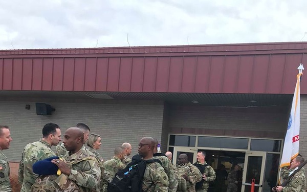 A group of U.S. Army soldiers in OCP (Operational Camouflage Pattern) uniforms are gathered outside a military building, appearing to be either departing or arriving at a military installation. Several soldiers are carrying backpacks and gear, and a military unit flag is visible on the right side. The scene takes place on a wet, overcast day on what appears to be a marked tarmac or parade area.