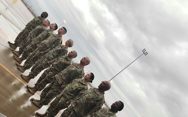 A group of approximately 10-12 military personnel in U.S. Army Operational Camouflage Pattern (OCP) uniforms are standing in a single-file line on a wet pavement surface. The image is rotated approximately 90 degrees, giving a tilted perspective. The setting appears to be a military installation or base, likely during or after rainfall.