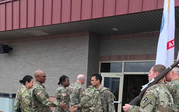 A group of U.S. Army soldiers in Operational Camouflage Pattern (OCP) uniforms are gathered outside a military building, engaged in a greeting or reception ceremony. Several soldiers are shaking hands, suggesting a welcome-back, departure, or change-of-command type event. Two soldiers on the right are standing at attention, with one carrying a unit guidon or flag.