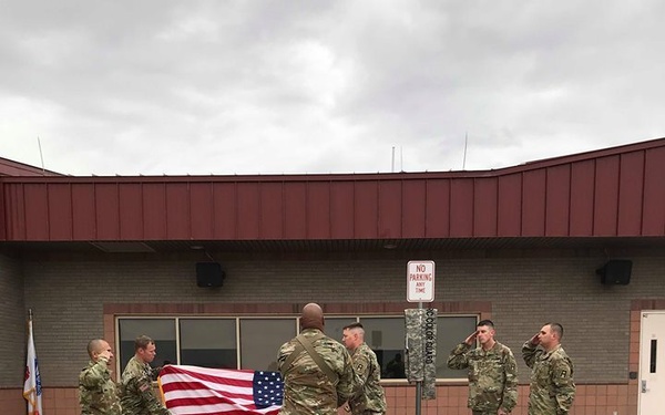 A group of U.S. Army soldiers in OCP (Operational Camouflage Pattern) uniforms are conducting a flag ceremony in a parking lot outside a military building. Three soldiers are handling and folding the American flag while two others stand at attention rendering a salute. The setting appears to be a military installation, likely during a retreat or colors ceremony.