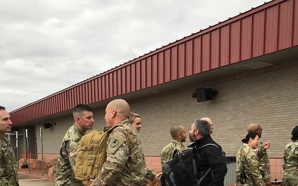 A group of military personnel in OCP (Operational Camouflage Pattern) uniforms are gathered on a wet tarmac or parade ground outside a brick building with a red metal roof. A civilian man wearing a black jacket and carrying a backpack is shaking hands with one of the uniformed soldiers. The scene appears to depict a greeting or reception ceremony, possibly a deployment departure or return.