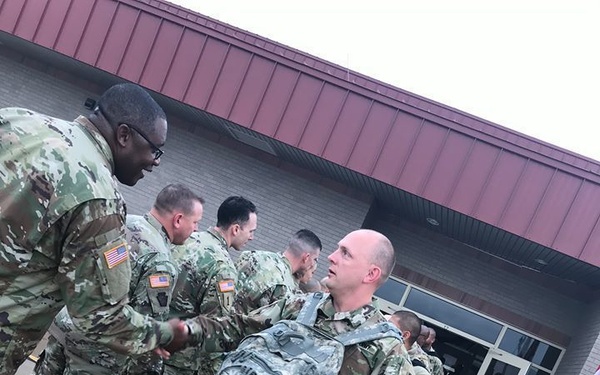 A group of U.S. Army soldiers in camouflage uniforms are gathered outside a military building, engaged in a handshake greeting or farewell ceremony. The central figure is a bald soldier carrying a large digital-pattern backpack and a hard-shell equipment case. The scene appears to be a deployment or redeployment event, with soldiers lined up near a building entrance.