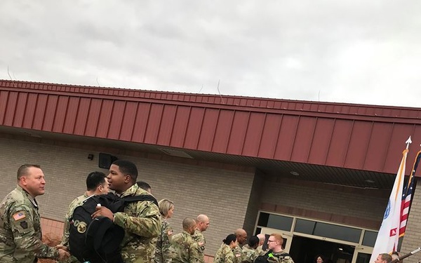 A group of U.S. Army soldiers in OCP (Operational Camouflage Pattern) uniforms are gathered outside a military building on a wet, overcast day. Several soldiers are embracing and greeting one another, suggesting a departure or return ceremony. A color guard soldier holds flags near the building entrance, and a 'NO PARKING' marking is visible on the wet pavement in the foreground.