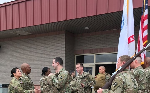A group of U.S. Army soldiers in OCP (Operational Camouflage Pattern) uniforms are gathered outside a military building, engaged in a greeting or farewell ceremony. Senior personnel on the left are shaking hands with soldiers who appear to be arriving or departing, with some carrying rucksacks. A color guard on the right holds what appears to be a unit guidon and the U.S. flag.