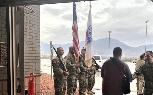 The image shows a military ceremony or reception taking place outside a brick building, viewed through an open doorway. Several soldiers in camouflage uniforms are holding flags, including the American flag and what appears to be a unit or organizational flag. A civilian in a red jacket is being greeted or escorted by military personnel.