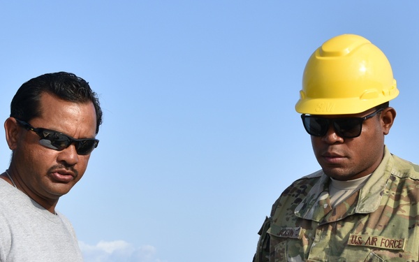 Two individuals are examining equipment at what appears to be a port or coastal construction site. One person is wearing U.S. Air Force OCP (Operational Camouflage Pattern) uniform with a yellow hard hat, while the other is in civilian attire (gray t-shirt) with sunglasses. Both are focused on a control panel or piece of equipment in the foreground.