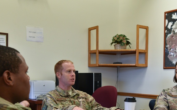 Three military personnel in OCP (Operational Camouflage Pattern) uniforms are seated around a wooden conference table in what appears to be an office setting. The individual in the center appears to be speaking while gesturing, suggesting a briefing or discussion is taking place. A tablet device and travel mug are visible on the table.
