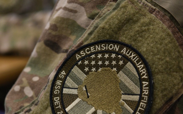 A close-up photograph of a military uniform patch worn on a camouflage (OCP/MultiCam) sleeve. The patch reads 'Ascension Auxiliary Airfield' and identifies the unit as '45 MSG DET 2' with geographic coordinates '7° 56'S 14° 22'W', corresponding to Ascension Island in the South Atlantic. The patch features overlapping American and British flag designs with a silhouette of Ascension Island in the center.