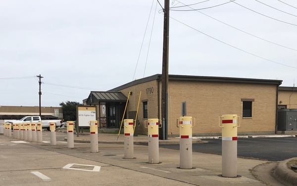The image shows a row of security bollards (yellow-topped with red reflective bands) lining a road in front of a tan brick building numbered 1790, which appears to be a military installation based on the visible signage referencing '47th Fighter Squadron'. The bollards serve as vehicle access control barriers, and the scene includes a stop sign, utility poles, and overhead power lines in an overcast setting.