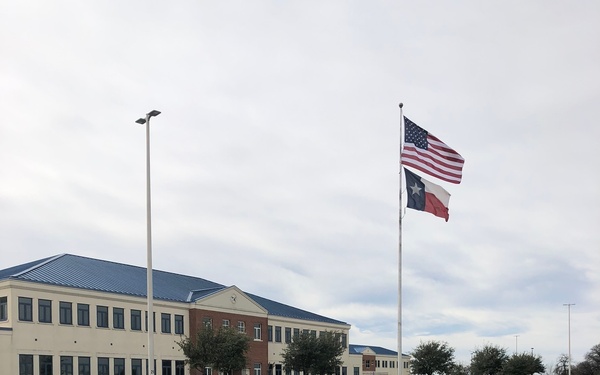 The image shows a large institutional or government building with a beige/cream exterior and blue metal roof, located in what appears to be Texas based on the Texas state flag flying alongside the U.S. flag on a tall flagpole. The facility features a well-maintained parking area with black and yellow bollards, manicured landscaping, and multiple light poles. The architectural style and security features suggest a government, military, or law enforcement facility.