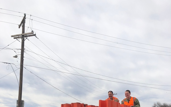 Four individuals wearing orange high-visibility safety vests are unloading large orange plastic barriers (likely water-filled traffic/security barriers) from a flatbed truck. Two individuals appear to be in military camouflage uniforms, suggesting this may be a military or government logistics operation. The scene takes place in what appears to be a military installation or industrial area.