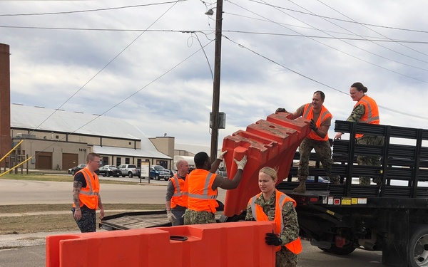 A group of military personnel wearing Navy camouflage uniforms and orange high-visibility safety vests are unloading large orange plastic traffic/water-fillable barriers from a flatbed truck onto a road. The operation appears to be taking place on a military installation or base, with industrial buildings visible in the background. Multiple personnel are coordinating the effort, with some on the truck bed passing barriers down to those on the ground.
