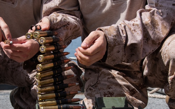 Military personnel in desert camouflage uniforms are handling a belt of linked ammunition. They appear to be loading or organizing rounds into an ammunition can. The setting appears to be an outdoor military environment with wooden pallets visible in the background.