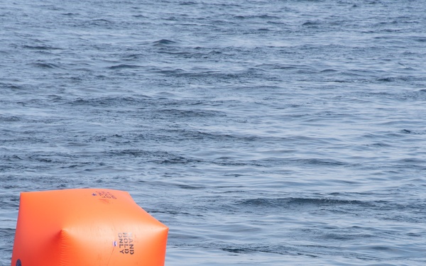An inflated orange cubic buoy is floating on open water with a slightly choppy blue-gray surface. The buoy appears to be a maritime marker or racing mark, with visible black text printed on its sides. It is tethered by a yellow line visible near its base.