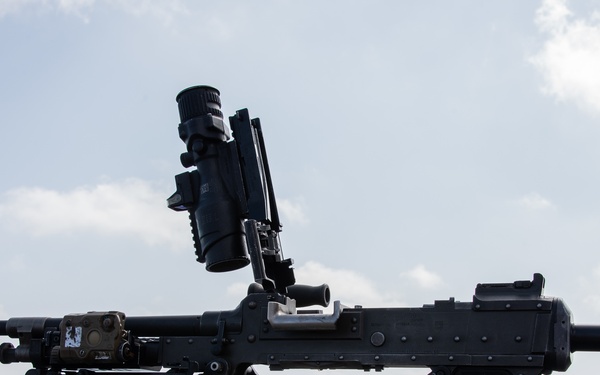 The image shows a mounted machine gun positioned on what appears to be the deck of a naval vessel. An ammunition box labeled '200 CARTRIDGES 7.62MM M13' is visible in the foreground with loose spent casings scattered on the deck, suggesting recent firing activity. The weapon is equipped with an optical scope and appears to be on a tripod or pintle mount.