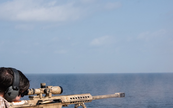 A military personnel member in desert camouflage uniform is firing a large-caliber sniper rifle from a wooden pallet platform over open ocean. The rifle appears to be a Barrett M82/M107 or similar .50 caliber anti-materiel rifle in a tan/desert finish, mounted on a bipod. Muzzle smoke is visible, indicating the weapon has just been discharged.