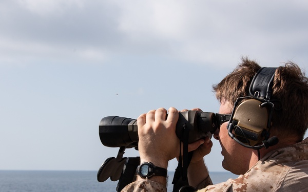 A military personnel member in desert camouflage (MARPAT) uniform is conducting maritime surveillance from the deck of a naval vessel using large binoculars. The individual is equipped with tactical hearing protection/communications headset and a tactical watch. The scene takes place over open ocean under partly cloudy skies.