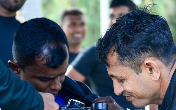 Two men in dark wetsuits or dive skins are closely examining and handling scuba diving equipment, including regulators, hoses, and gauges. A third person is visible smiling in the background, suggesting a group diving activity or training session. The setting appears to be outdoors, likely a dive site preparation area.