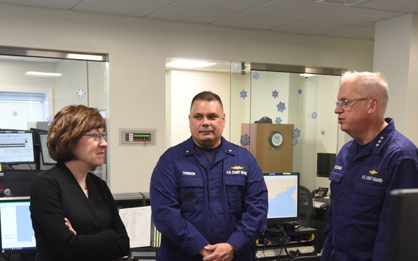 A civilian woman in a black blazer is meeting with U.S. Coast Guard personnel in what appears to be an operations or command center. Several uniformed Coast Guard members are present, including officers with visible rank insignia and name tapes. The setting includes multiple computer workstations and holiday snowflake decorations on the glass partitions.