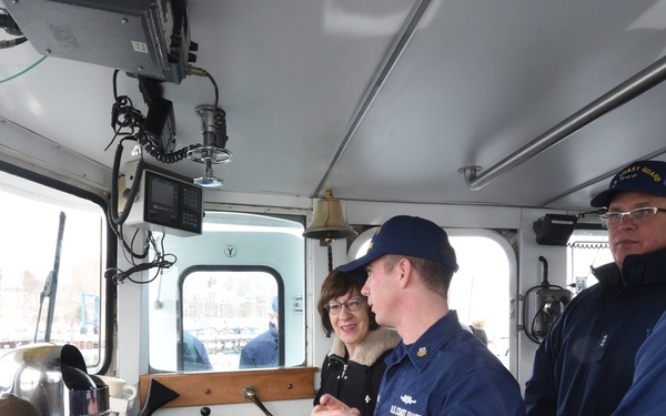 The image shows the interior of a U.S. Coast Guard vessel's bridge or pilothouse, where several Coast Guard personnel in navy blue uniforms are gathered alongside a civilian woman. A Coast Guard member appears to be explaining or demonstrating the vessel's controls and navigation equipment. The setting suggests an official tour or briefing aboard the vessel.