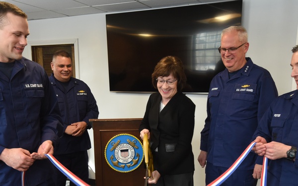 A ribbon-cutting ceremony is taking place at a U.S. Coast Guard facility. A civilian woman in a black jacket is cutting a red, white, and blue ribbon using gold scissors, flanked by U.S. Coast Guard personnel in navy blue uniforms. A podium bearing the U.S. Coast Guard seal is visible in the background.