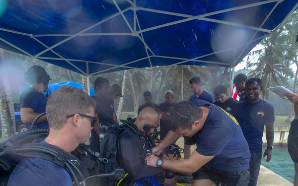 A group of individuals are gathered under a blue canopy tent in what appears to be a rainy outdoor setting near a waterfront or dive site. Several people are wearing or being fitted with scuba diving equipment including BCDs, tanks, regulators, and fins. The scene suggests a dive training or preparation session, likely military or organized dive team based on the matching dark uniforms worn by most participants.