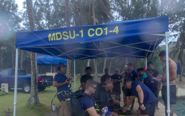 A group of military divers from MDSU-1 CO1-4 (Mobile Diving and Salvage Unit One) are gathered under a blue canopy tent near a waterfront, preparing for a dive operation. Personnel are seen donning scuba gear including tanks and BCDs, while equipment cases and yellow air hoses are staged on the concrete dock. The setting appears to be a tropical location with palm trees and calm water visible in the background.