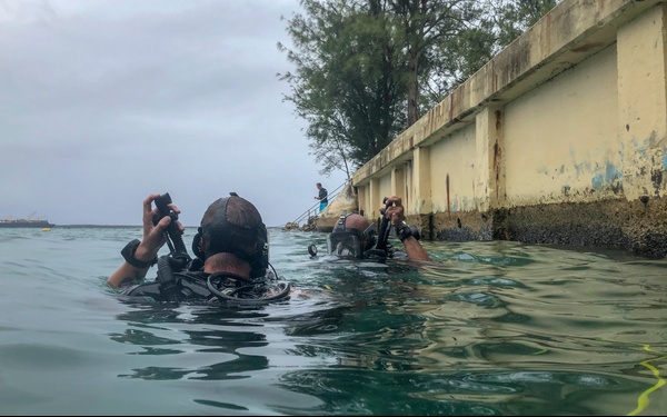 Two scuba divers are visible at the water surface near a weathered concrete seawall, with a third person standing on steps leading down to the water in the background. The divers are fully equipped with diving gear including tanks, wetsuits, and regulators. The scene appears to be a shore dive entry or exit point under overcast skies.