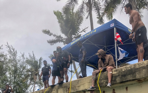 A scuba diver in full gear is in the water, holding a camera (appears to be a GoPro setup), photographing or filming a concrete seawall. Above on the seawall, a group of people in dive gear and casual clothing are gathered under a blue canopy tent marked 'MDSU-1 CO1-4', with yellow tether/safety lines running from the wall down to the water. The setting appears to be a military or official dive operation near a tropical coastline.