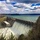 A large concrete gravity dam is shown releasing water through multiple spillway gates, creating a dramatic white-water discharge at its base. A substantial reservoir with turquoise-green water stretches behind the dam, surrounded by forested hills. The image is taken from an elevated vantage point on a partly cloudy spring day, with bare deciduous trees visible in the foreground.