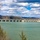 The image shows a large concrete dam spanning across a wide body of water with a green-tinted reservoir in the foreground. The dam features multiple spillway gates and a control structure on the right side, set against a backdrop of wooded hills and a partly cloudy sky. The scene appears to be captured during late winter or early spring based on the bare deciduous trees.