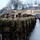 A formation of U.S. Marines in MARPAT digital camouflage uniforms and covers stands at attention during what appears to be a formal military parade or ceremony. The Marines are viewed from behind, holding rifles, facing toward a large crowd of spectators in bleachers. Flags including the Maryland state flag and the U.S. flag are visible in the background.