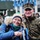A civilian woman and a military serviceman pose together smiling at what appears to be a public military event or parade. The woman holds small Estonian flags (blue, black, and white tricolor) and has camouflage face paint applied. In the background, other military personnel including a soldier with a US flag patch are visible, along with military equipment.
