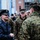 A senior U.S. Air Force officer in a dress blue overcoat and service cap is interacting with U.S. Marines in MARPAT camouflage uniforms in an urban outdoor setting. The Air Force officer appears to be greeting or conversing with the Marines, possibly at a public event or ceremony. The setting appears to be a city street or plaza with buildings visible in the background.