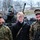 Two soldiers in digital camouflage uniforms (consistent with US Marine Corps MARPAT pattern) pose with a civilian young man at what appears to be a military public display event. In the background, a military armored vehicle is visible with a child sitting in it, surrounded by a crowd of spectators. A monument inscription partially reading 'Eesti Vabadussõda' (Estonian War of Independence) is visible in the background, suggesting this event takes place in Tallinn, Estonia.