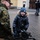 A smiling child in winter clothing interacts with a military serviceman in digital camouflage uniform during what appears to be a public military demonstration or outreach event. The child is kneeling behind a mounted military weapon system, likely a machine gun on a bipod, and appears excited and engaged. The setting is an outdoor cobblestone plaza with European-style architecture visible in the background.