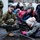 A U.S. Marine in MARPAT camouflage uniform is supervising a young girl as she looks through the scope of a large machine gun mounted on a bipod, likely at a public military demonstration or open day event. An adult civilian man in a flat cap and winter jacket assists or observes alongside her. The setting appears to be an outdoor urban plaza in cold weather, with other military personnel and civilians visible in the background.